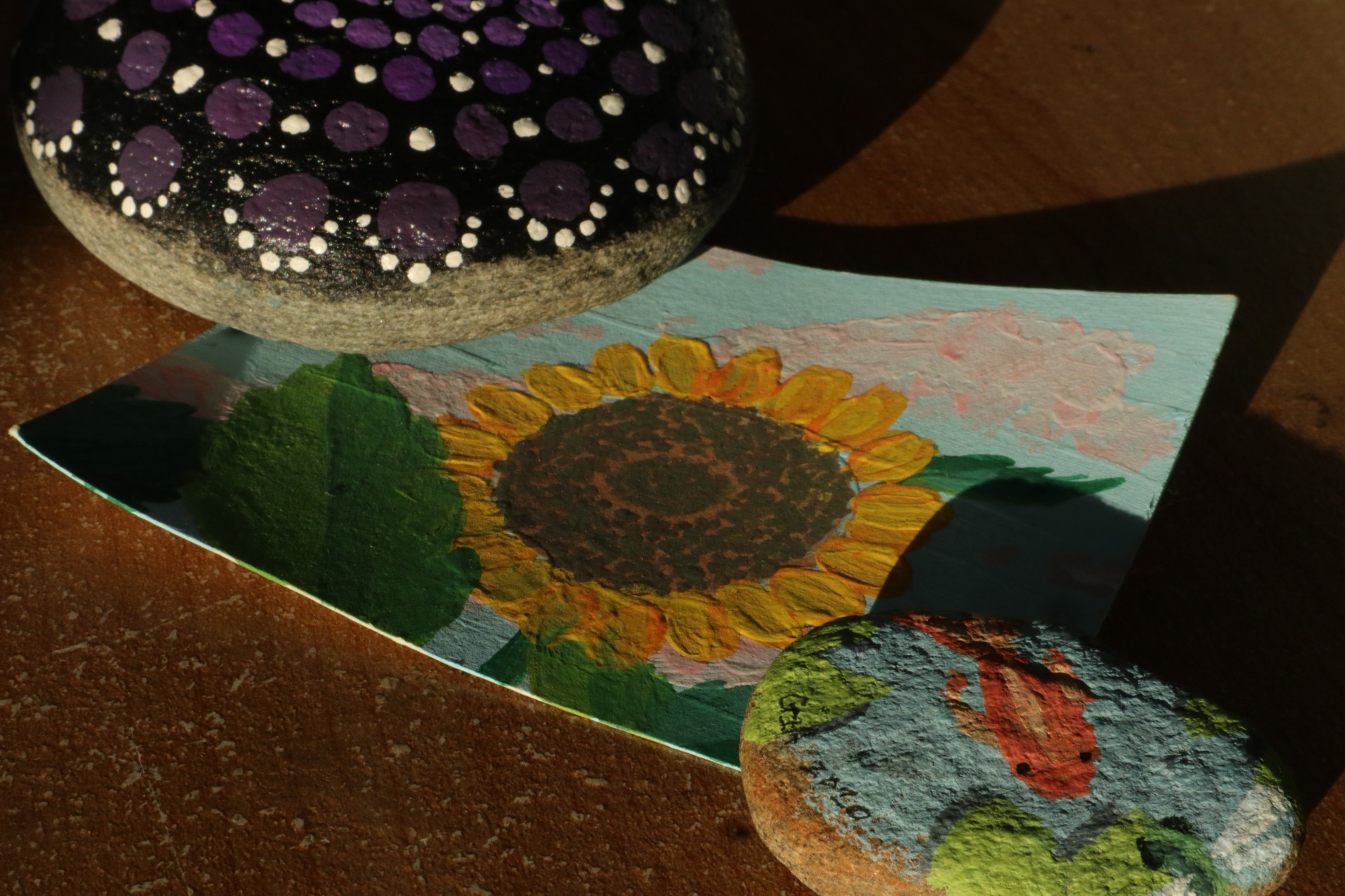 One large rock with a black background with symmetric gradient purple and white dots and a smaller rock with a painted kio fish pond holding down an acrylic sunflower painting